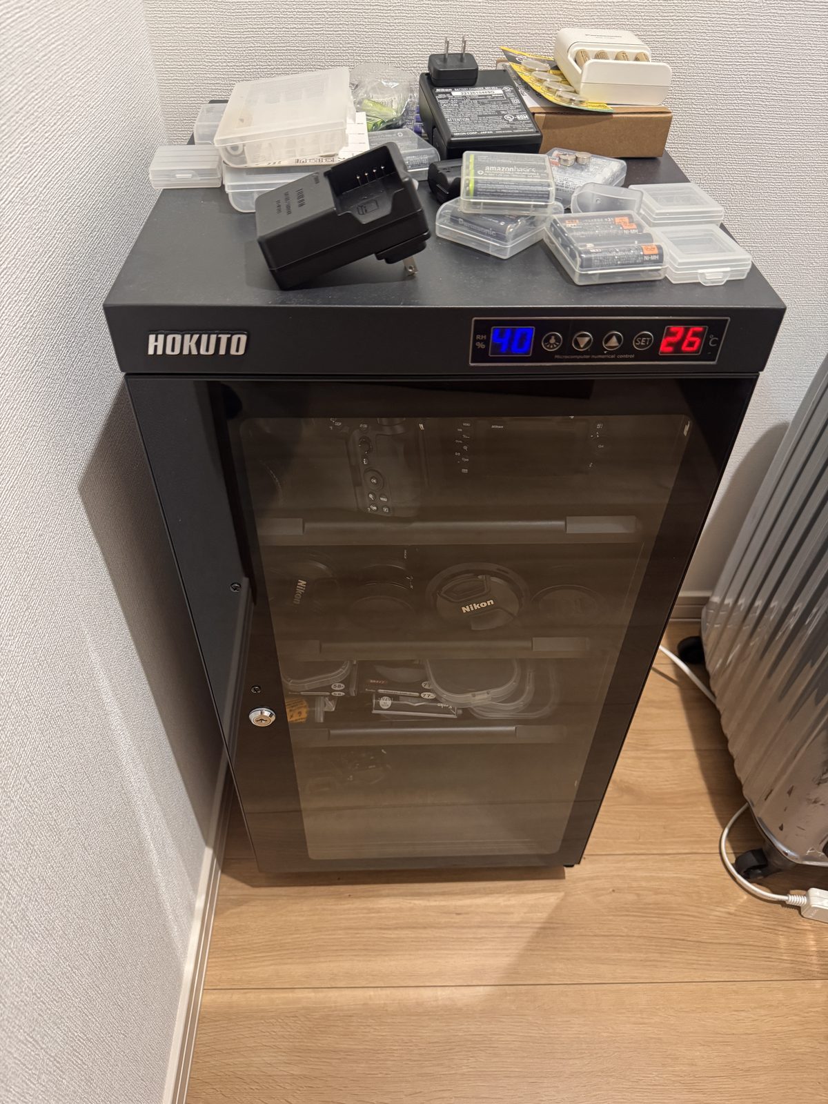 部屋の隅に置いた黒いHOKUTOの防湿庫。扉はガラスで、上部のパネルに湿度と温度の数字が表示されている