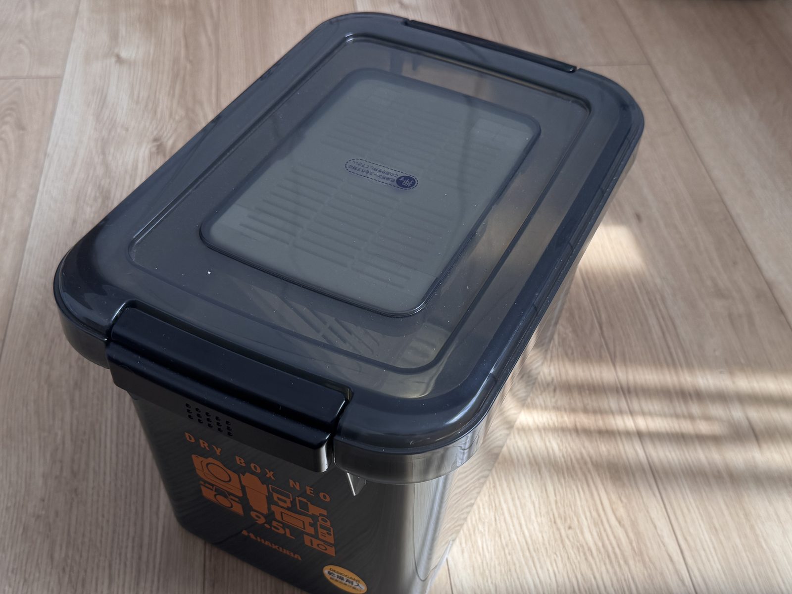 床に置いたハクバのドライボックスNEO 9.5L。スモークの半透明で、側面に DRY BOX NEO 9.5L と印刷されている