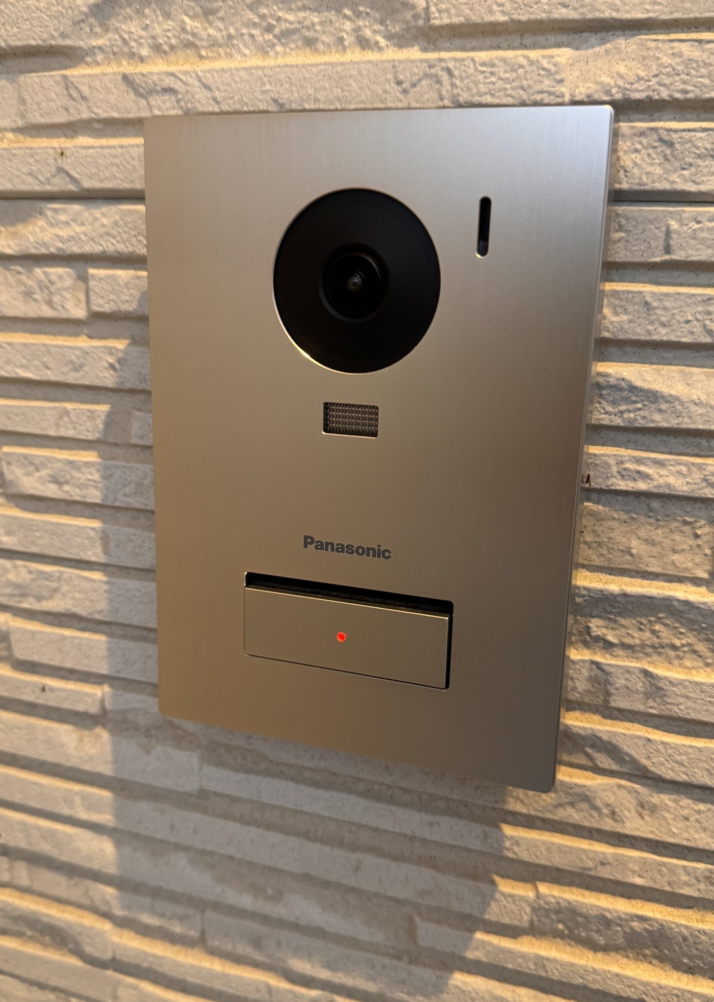 玄関まわりの石積みの壁に取り付けられた、銀色のパナソニックの玄関子機。上に丸いカメラ、下に赤いランプの付いた呼び出しボタンがある