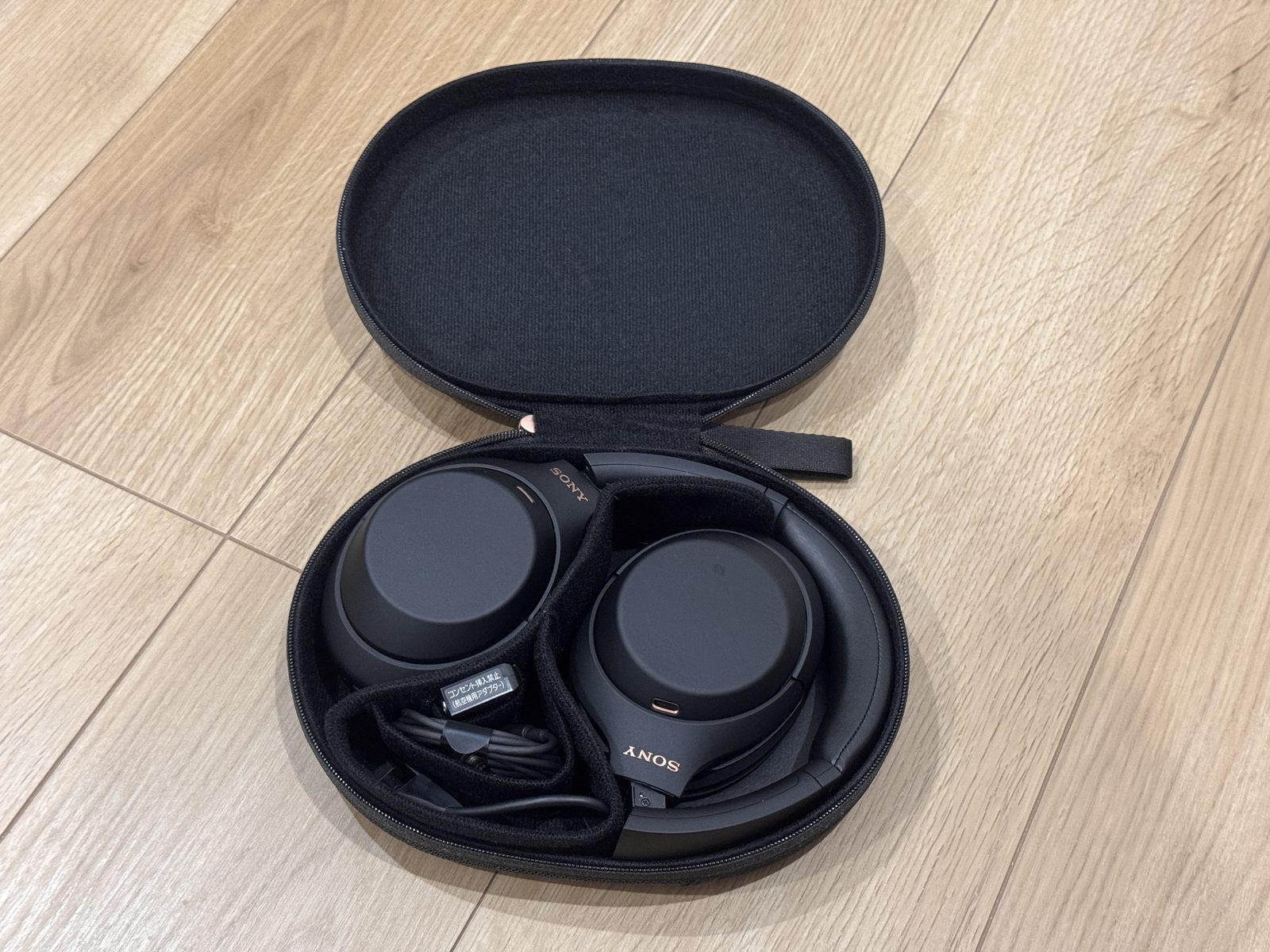 開いたハードケースに入った黒いSONY WH-1000XM4。左側に充電ケーブルと航空機用アダプターが収まっている